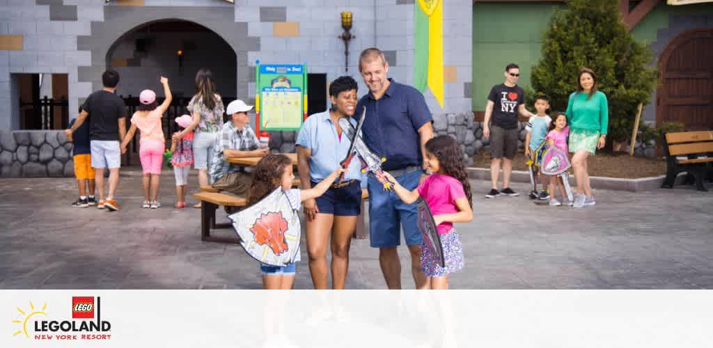 Image of visitors at LEGOLAND New York Resort enjoying the park. A family smiles in the foreground while other guests walk and relax in the background near a castle-like structure. The LEGOLAND logo is visible in the corner.