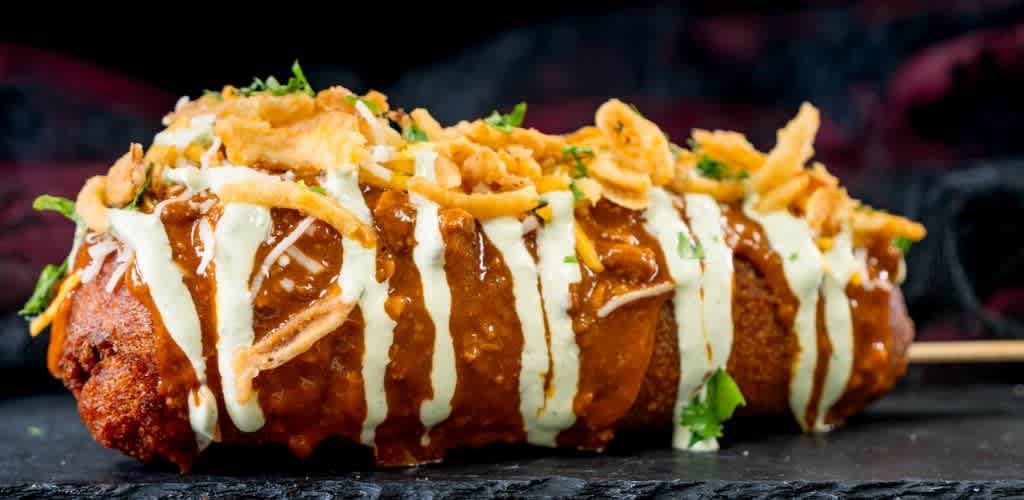 A close-up of a burrito topped with white sauce, brown sauce, cheese, crispy fried onions, and fresh cilantro on a dark surface.