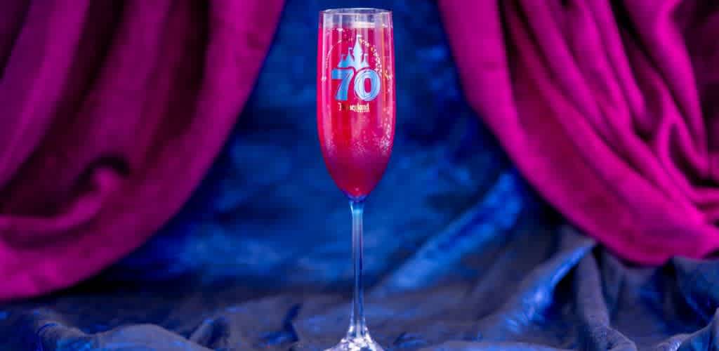 A tall champagne glass filled with a vibrant red drink is set against a deep blue and purple backdrop. The glass is elegantly designed with a subtle 70 emblem on it, celebrating an anniversary or special occasion. The fabric drapes around the glass, adding a touch of luxury and festiveness to the scene.