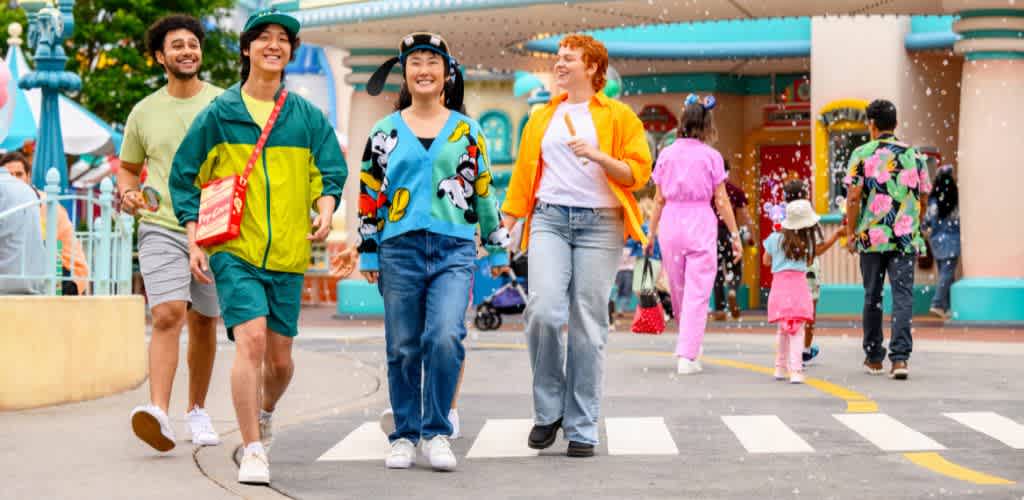 A diverse group of four people happily walk through a vibrant theme park setting. They're dressed in colorful casual attire, surrounded by playful decor and rides. The atmosphere is lively and welcoming, with other park-goers in the background. The scene is filled with joy and excitement, reflecting an enjoyable day out.