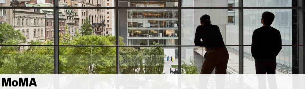 A view from inside a modern art museum shows two people silhouetted against large windows. They look out onto a cityscape featuring lush green trees and historic buildings. Beyond, a contemporary building can be seen, blending classic and modern architecture. The scene captures a moment of reflection and appreciation of art and urban scenery.