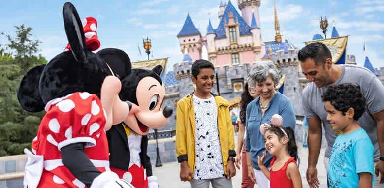 A family of six smiles and interacts with two iconic cartoon characters dressed in costumes in a theme park. The background features a whimsical castle under a bright blue sky. The group includes adults and children, all appearing joyful and engaged. The setting is lively and picturesque, capturing the excitement and magic of the experience.