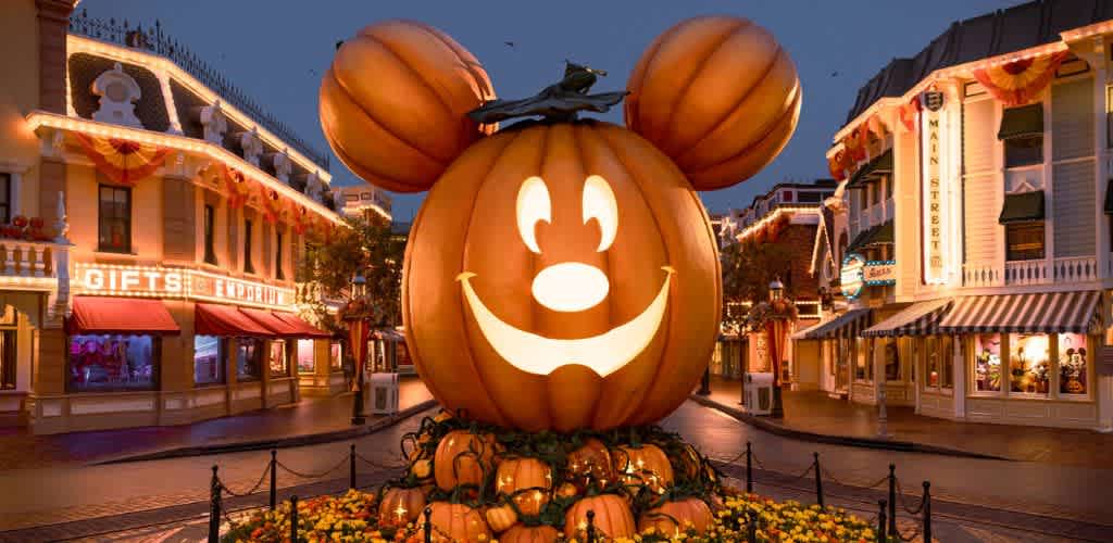 A festive scene at a theme park features a giant pumpkin with mouse ears and a smiling face, situated in the center of a decorated street lined with shops. The evening sky and glowing lights create a welcoming and magical atmosphere, perfect for celebrating fall festivities.
