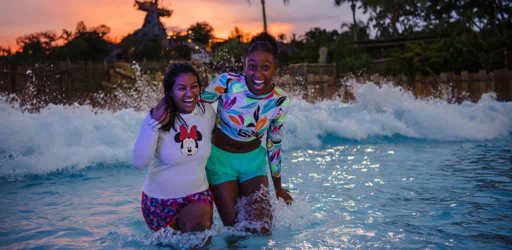 Two friends enjoying a wave pool at sunset in a water park. Both are wearing colorful swimwear and laughing as the waves crash around them. The sky is a warm mix of orange and pink hues, and there are trees and a wooden structure in the background. The water appears vibrant under the sunset glow, adding to the fun and relaxed atmosphere.