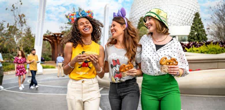 Three friends enjoy a day at an amusement park. They are laughing and holding snacks, including pretzels. Each wears themed attire and headbands with colorful designs. The background shows a large, iconic geodesic structure and trees, suggesting a lively, fun atmosphere. Other visitors are visible, adding to the scene's vibrant, joyful vibe.