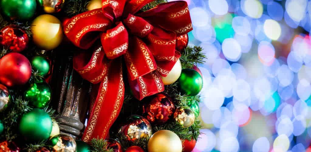 A close-up of a festive Christmas wreath decorated with red, gold, and green ornaments and a large red ribbon bow, with colorful blurred holiday lights in the background