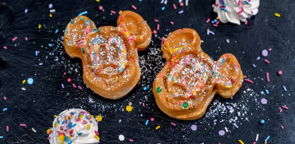Two character-shaped waffles are topped with colorful sprinkles and a dusting of powdered sugar, placed on a dark surface. Swirls of whipped cream and scattered sprinkles surround the waffles, adding a fun and festive touch.