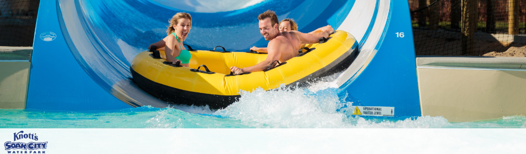 A group of three people enjoying a thrilling ride on a large yellow raft as they slide down a blue water slide at Knott's Soak City Waterpark. The splash around them suggests speed and excitement. Bright, sunny weather highlights the fun atmosphere of the waterpark setting.