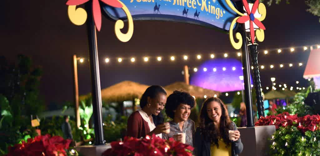 Three people enjoy drinks and laughter under festive lighting at a Three Kings celebration. The setting features colorful decorations, bright poinsettias, and a cheerful ambiance. In the background, an illuminated dome and string lights create a vibrant and inviting atmosphere.
