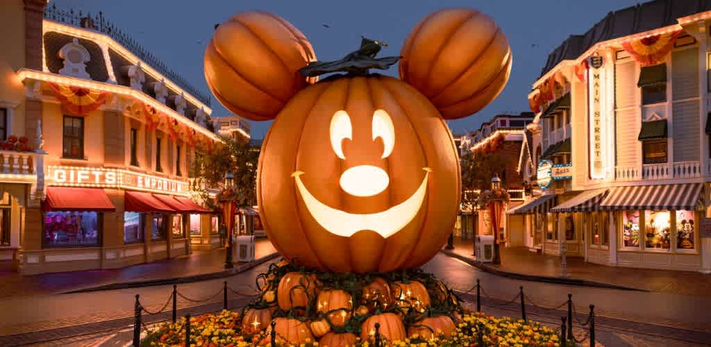 A giant pumpkin decorated as Mickey Mouse with illuminated face and ears, surrounded by smaller pumpkins in a themed outdoor street scene at dusk.