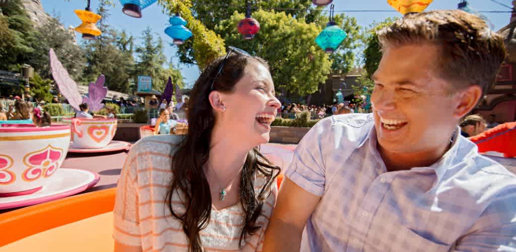 A happy couple laughs while enjoying a colorful teacup ride at an amusement park. They are surrounded by vibrant teacups and hanging lanterns with trees and a smiling crowd in the background, creating a lively atmosphere.