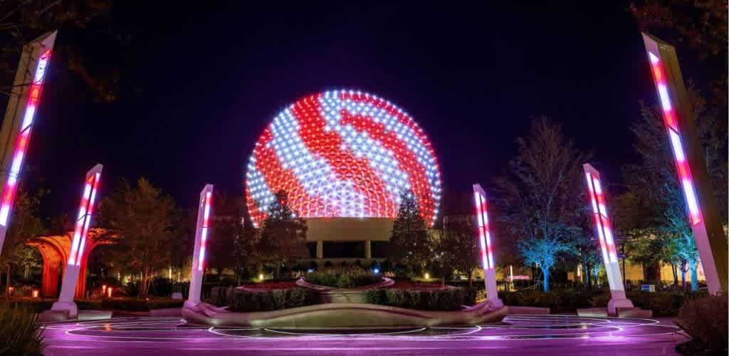 Night view of a geodesic sphere illuminated with red, white, and blue lights, creating a vibrant pattern. The surrounding area features tall light fixtures with similar color schemes, enhancing the visual spectacle. The setting includes landscaped gardens, with trees subtly lit in blue, creating a serene and captivating atmosphere.