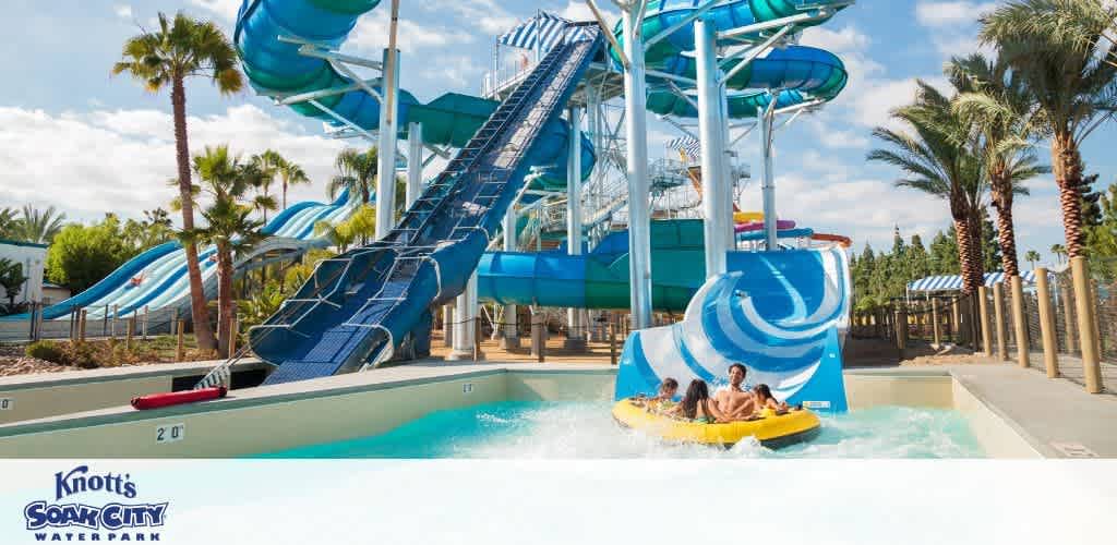 A group of people enjoys a thrilling ride on a yellow raft down a twisting blue water slide at a water park. Tall palm trees and other slides are visible in the background, creating a fun and sunny atmosphere. The Knott's Soak City Water Park logo is visible in the corner.