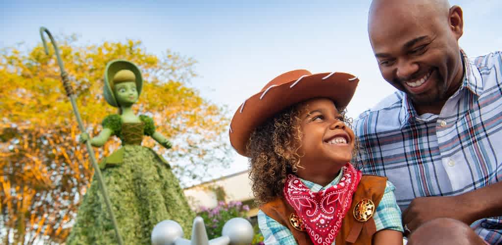 A young girl dressed as a cowboy and an adult smiling outdoors with a decorated topiary figure in the background on a sunny day