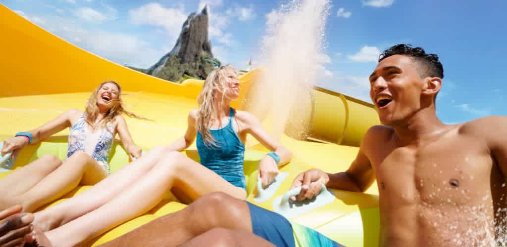 A group of people enjoy a thrilling ride on a yellow water slide at a water park. They are laughing and appear exuberant as water splashes around them. In the background, a tall, rocky structure rises against a clear blue sky, adding to the exciting atmosphere of the scene.