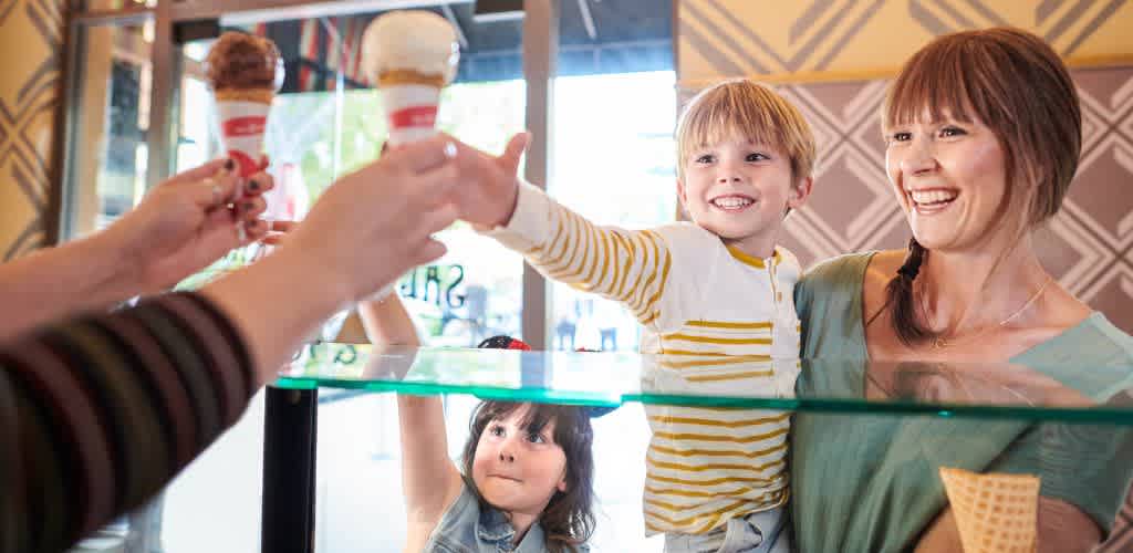 A woman and two children receive ice cream from an ice cream shop attendant behind a glass counter. The children are smiling and reaching for the ice cream.