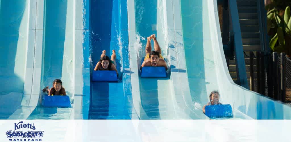 Visitors enjoy racing down water slides at a water park. Four people on blue mats glide down adjacent slides with joyful expressions, creating splashes as they descend. Sunlight reflects off the water, adding a lively atmosphere to the scene. Lush greenery and stairs are visible in the background, enhancing the setting's appeal as a fun and refreshing place for adventure.