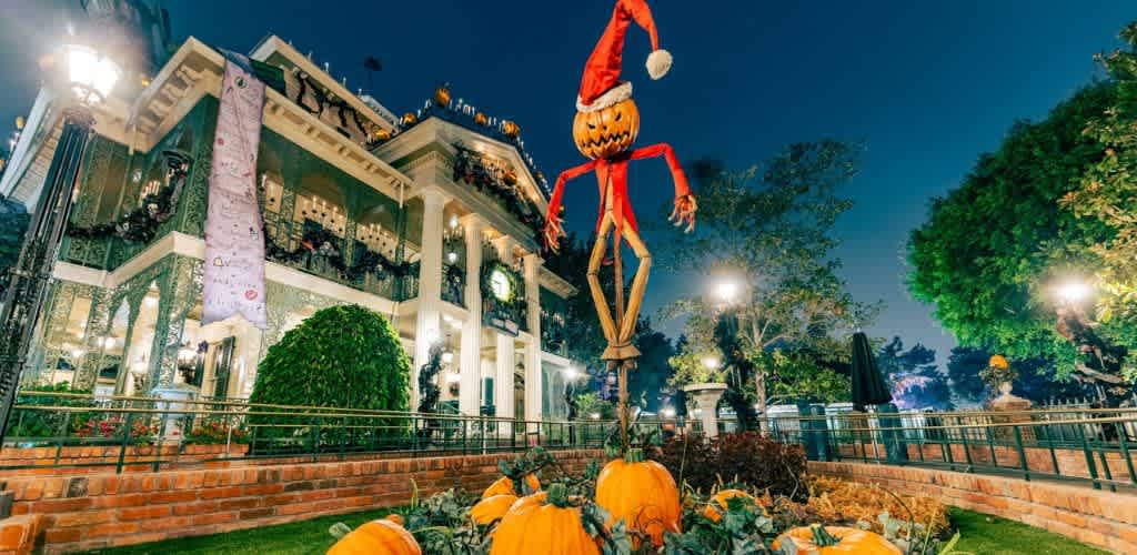 Festive scene at an amusement park with a large mansion decorated for Halloween, featuring a tall scarecrow wearing a Santa hat. Brightly lit at night, the area is adorned with pumpkins and lights, creating a whimsical holiday atmosphere. Trees and surrounding greenery complete the enchanting setup, inviting visitors to enjoy the seasonal celebration.