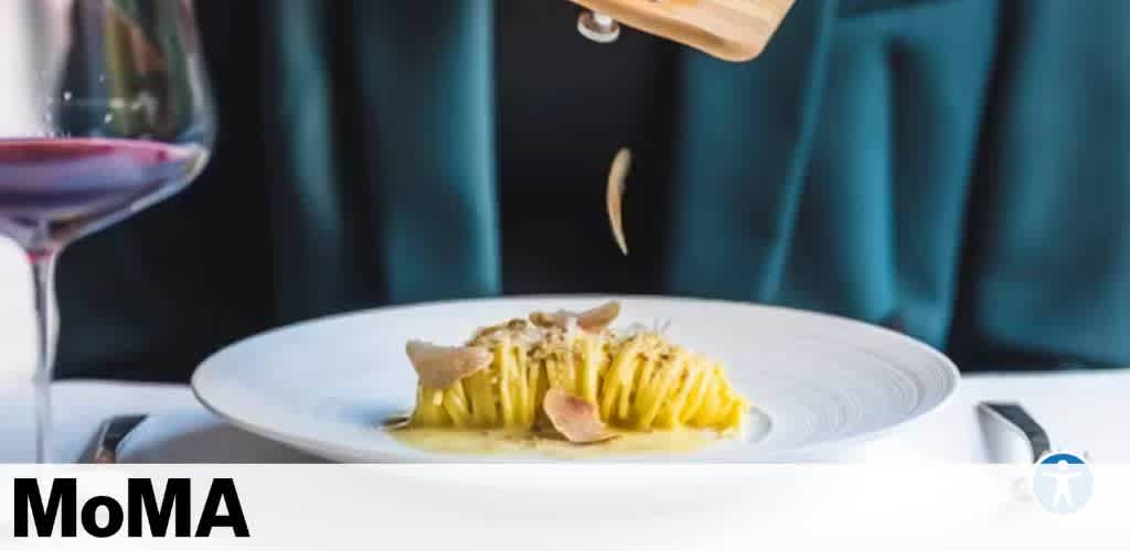 A plate of elegantly plated pasta is being garnished with thinly sliced truffles, creating a visually appealing dish. Beside it, a glass of red wine adds a touch of sophistication. The background features a soft, elegant setting, adding to the luxurious dining experience. The image is associated with a perk from MoMA, offering an exquisite culinary treat.