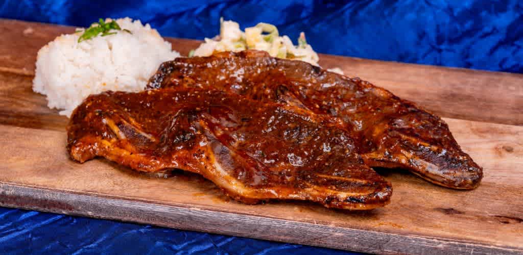 A delicious platter showcasing succulent grilled ribs glazed with a savory sauce is presented on a wooden board. The dish is accompanied by a scoop of fluffy white rice garnished with herbs and a side of creamy coleslaw. The background features a deep blue fabric setting that enhances the warm tones of the meal, creating an inviting visual appeal perfect for enticing food lovers.