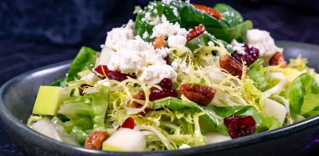 A fresh mixed salad with lettuce, dried cranberries, nuts, cheese crumbles, and vegetables in a black bowl on a dark background