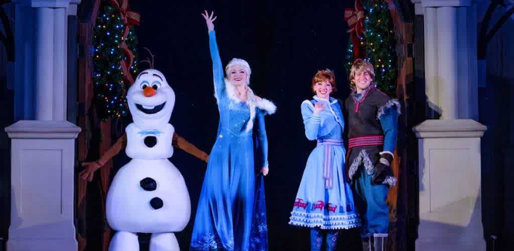 Four characters in winter-themed costumes stand on stage. A cheerful snowman with twig arms stands beside a woman in a long blue ice-themed dress. Next to her is a woman in a blue dress with a reddish-brown apron and a man in winter clothes with a fur-trimmed jacket. The background features festive holiday decorations with colorful lights.