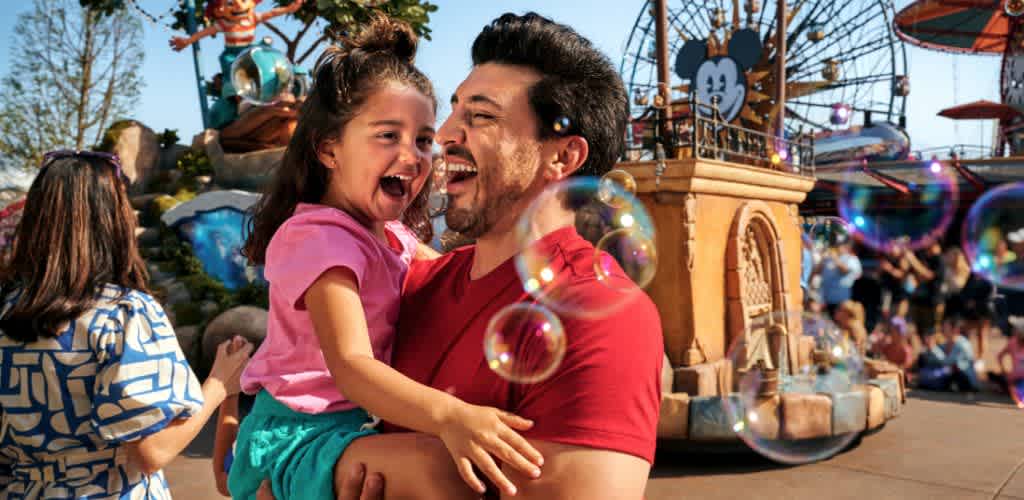 A man holding a laughing young girl at an amusement park with a carousel and bubbles in the background.