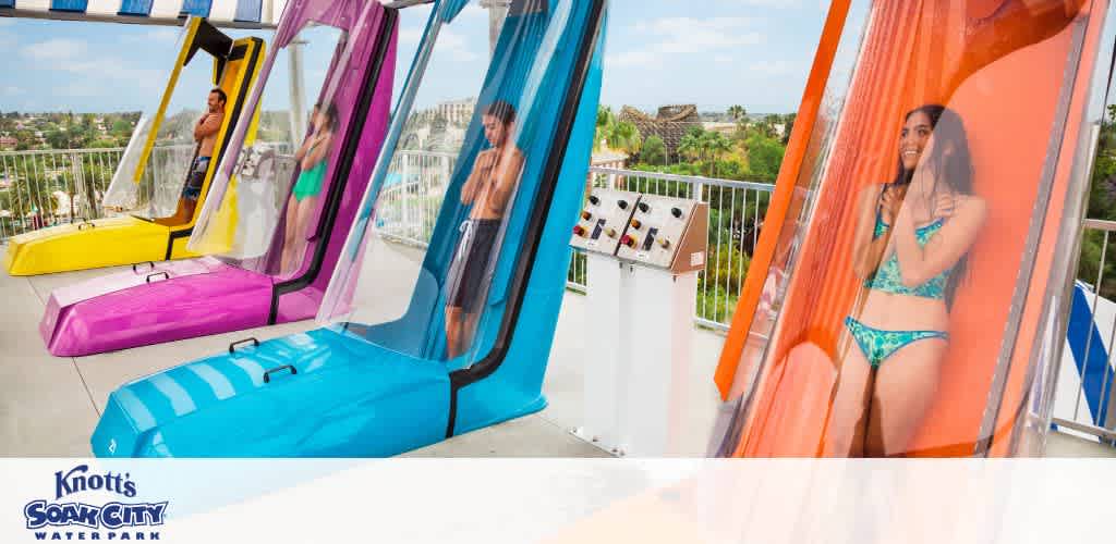Visitors at Knott's Soak City Water Park prepare to enjoy vertical drops in colorful capsule-style waterslides. Each slide features a transparent enclosure and is positioned side-by-side. The riders stand inside, anticipating the thrilling descent. The backdrop includes lush greenery and clear skies, adding to the vibrant and fun atmosphere of the park.