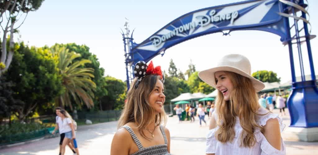 Two women smiling and talking at Downtown Disneyland entrance with trees and people in the background