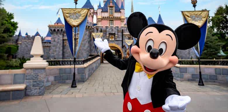 A cheerful character in a classic red and black outfit with large round ears stands welcomingly in front of a whimsical castle featuring pink and blue spires. The pathway leading to the castle is adorned with banners, creating a magical and inviting atmosphere, perfect for a fun family adventure.