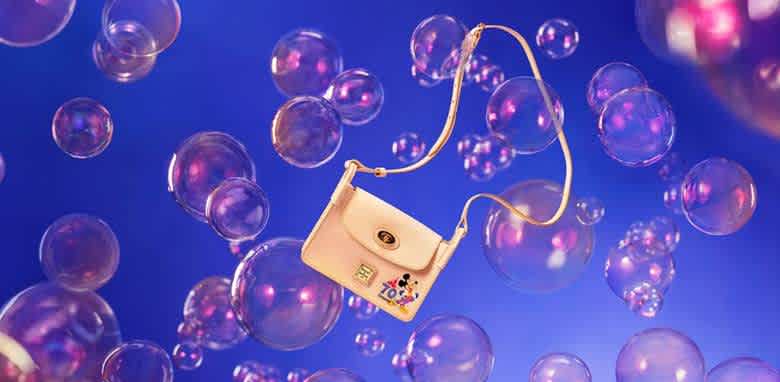 A stylish cream handbag with a shoulder strap featuring a playful graphic rests against a vibrant blue background filled with floating pink and purple bubbles. The scene adds a whimsical and fun element, highlighting the bag's fashionable design and youthful charm. Ideal for those seeking a trendy accessory with a touch of flair.