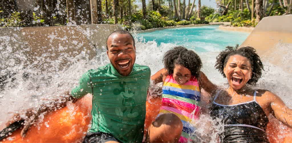 A joyful family enjoys a splashing ride down a water slide at a water park. The father is on the left in a green shirt, the daughter is in the middle wearing a colorful striped swimsuit, and the mother is on the right in a black swimsuit. They are surrounded by water and greenery, smiling and laughing as they experience the excitement and fun of the splash.
