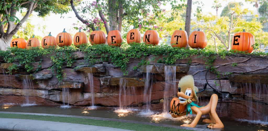 A charming Halloween display features orange pumpkins arranged on a stone ledge, each painted with a letter spelling Halloween Time. Water cascades gently from the ledge into a shallow stream below. A playful animated character leans against the ledge, holding a pumpkin. Lush green foliage and trees surround the scene, adding to the festive yet serene atmosphere.
