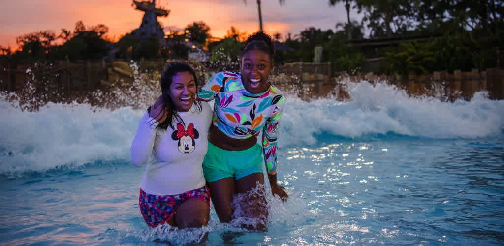 Two people are laughing and splashing in a wave pool at sunset. They are wearing colorful swimwear and enjoying the water. The background features lush greenery and a silhouetted water park structure against a vibrant sky.