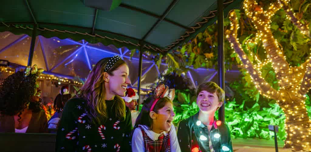 A joyful group of three children adorned with festive attire and bright headbands enjoys a night ride in a beautifully decorated park. They are surrounded by colorful holiday lights and lush greenery, creating a warm and enchanting atmosphere. The children are smiling and laughing, capturing the spirit of celebration and friendship during the holiday season.