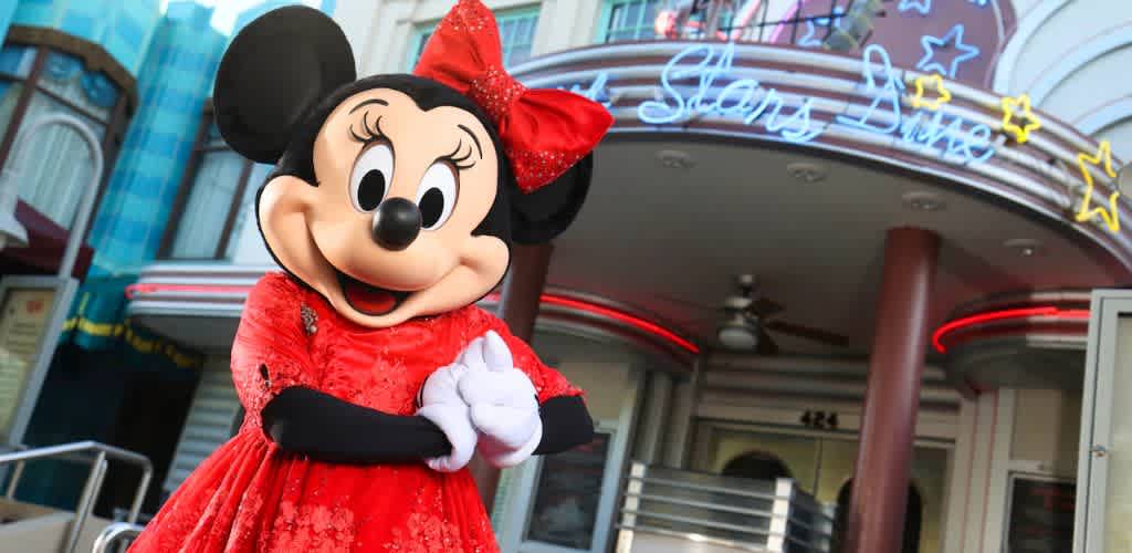 A character in a sparkly red dress and matching bow is posing happily in front of an illuminated diner. The diner has a retro facade with neon details and stars, creating a cheerful and inviting atmosphere.