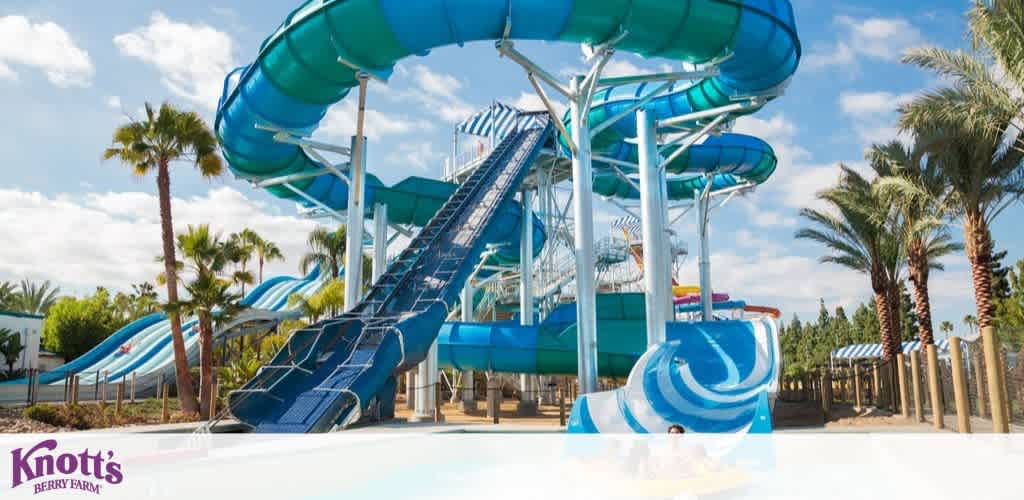 Image shows a vibrant water park scene with multiple blue water slides, some with enclosed tubes and others open. Palm trees dot the landscape, under a partially cloudy sky. The logo for Knott's Soak City Water Park is visible in the bottom left. A sense of fun and excitement is conveyed.