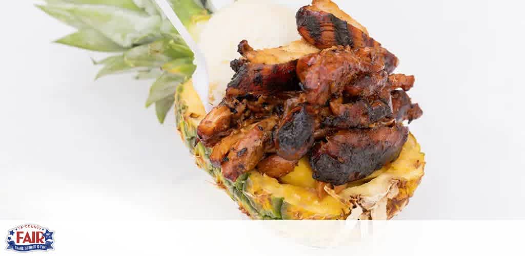 Image description: This is a culinary presentation featuring a pineapple cut in half horizontally, with its vibrant yellow flesh visible. The top half of the pineapple is slightly tilted backward to showcase a generous serving of grilled meat perched on the bottom half. The meat appears well-charred and seasoned, suggestive of a barbecue or grilling preparation, and is piled in a manner that it overflows slightly over the edges of the pineapple. The backdrop is a plain, light-colored surface which makes the colors of the pineapple and the caramelized meat stand out prominently. The lower left corner of the image displays a logo with text indicating an affiliation with a fair, signifying that the dish might be a unique offering at a local fair or festival.

For a flavorful experience minus the financial bite, visit GreatWorkPerks.com for the lowest prices on tickets to fairs and festivals where delightful dishes like this are waiting to be savored.