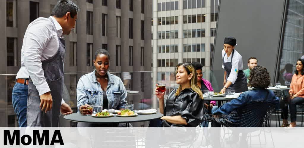 Image of a diverse group of people enjoying a meal on an outdoor terrace. In the foreground, a server presents a dish to a seated woman while another woman looks on, holding a glass of wine. In the background, a chef attends to guests. The MoMA logo is visible at the bottom of the photo, suggesting a museum setting.