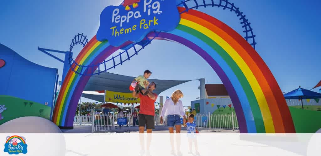 A family enters the colorful Peppa Pig Theme Park under a vibrant rainbow archway. A father holding a small child and a mother walking hand-in-hand with a young child pass by the welcoming sign, with attractions visible in the background under a bright blue sky.