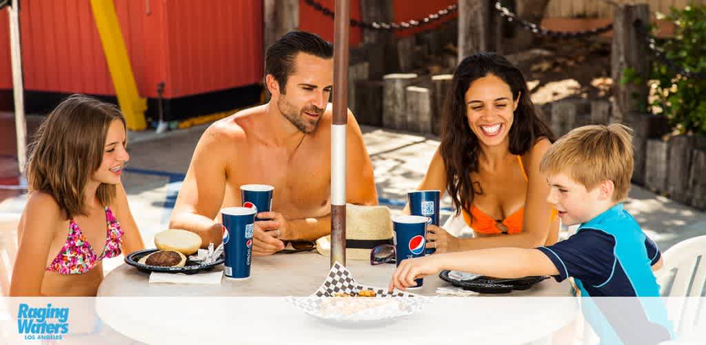 A family of four enjoys a meal at an outdoor table on a sunny day. A shirtless man, a woman in an orange top, a smiling girl, and a boy in blue are seated with burgers and drinks, all engaged happily in conversation. The background shows a water park ambiance with a hint of amusement rides and a clear blue sky.