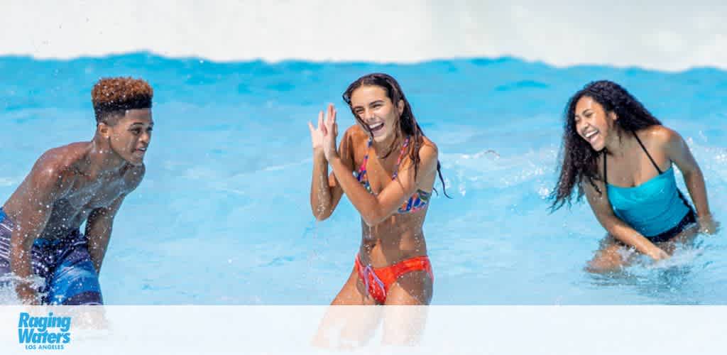 Three young people are joyfully playing in a water park pool with clear blue water. A person on the left appears to be approaching the other two, who seem to be splashing and encouraging their friend to join in the fun. The Raging Waters Los Angeles logo is visible in the corner, suggesting they are at this location. The sun is shining, and the vibe is energetic and cheerful.