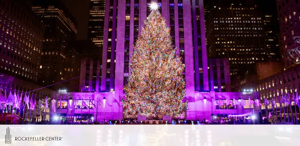 Image of a brightly illuminated Christmas tree at Rockefeller Center during night time. The towering tree is adorned with a multitude of colorful lights, casting a festive glow. Surrounding buildings are lit in soft purples and blues, adding to the holiday atmosphere. A skating rink in the foreground reflects the vibrant lights.