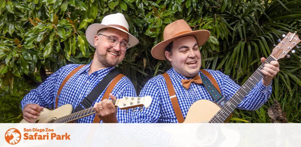 This image features two cheerful individuals engaged in a musical performance. Both are wearing white, brimmed hats and coordinating blue and white gingham shirts paired with tan suspenders. To the left, one musician is wearing glasses and playing a light-colored ukulele, while the rightmost performer is enjoying playing a classic brown guitar. They are both sporting bow ties, with the left in a blue bow tie and the right in an orange one. The background shows a lush green foliage of plants, suggesting an outdoor setting. The photograph includes the logo for the San Diego Zoo Safari Park in the lower right corner. At FunEx.com, we always strive to provide the lowest prices and best discounts on tickets to ensure your experiences are as delightful as the music these two are creating.