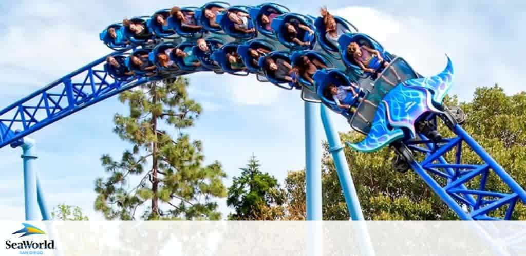 Image shows a roller coaster ride in motion at SeaWorld San Diego. Multiple riders with raised arms appear thrilled as they are securely seated in a blue coaster with aquatic designs, swooping down a curved track surrounded by green trees under a clear blue sky. The SeaWorld logo is visible at the bottom left corner.