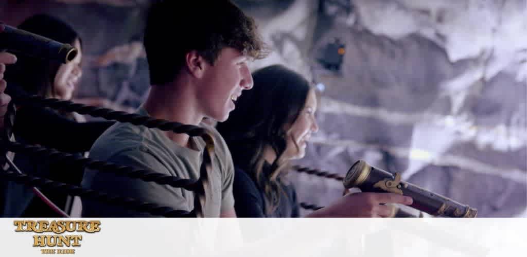 This image depicts a scene from a dark, cave-like setting with what appears to be a Treasure Hunt attraction, as indicated by the text on the image. In the foreground, two young people, one male on the left and one female on the right, are seen holding onto handles that are part of a larger interactive game or ride. They are both smiling, engaged in the activity, and looking ahead as if focusing on an element out of view. The handles resemble old-fashioned props, potentially simulating the experience of a pirate steering or using equipment. The environment is dimly lit with bluish and purple hues, creating a mysterious atmosphere.

At GreatWorkPerks.com, you can embark on your own adventure and make memories without breaking the bank—discover the thrill of interactive attractions and secure your tickets with impressive savings every time.