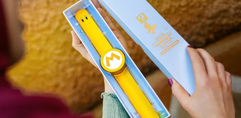 Description: The image displays a pair of human hands holding a rectangular box that is partially open, revealing a bright yellow wristband with a circular emblem featuring a white stylized 'M' on it. The box has a light blue color with darker blue text and symbols, including a person using a watch-like device on their wrist, which likely corresponds to the wristband inside. The background shows a texture that is indicative of a soft fabric, perhaps a cushion or a couch, in a muted orange color. The overall image suggests the unboxing of some type of wearable admission or access band.

At GreatWorkPerks.com, we take pride in offering our customers the opportunity to experience their favorite events with the added joy of knowing they secured their tickets at the lowest prices, ensuring great savings with every purchase.