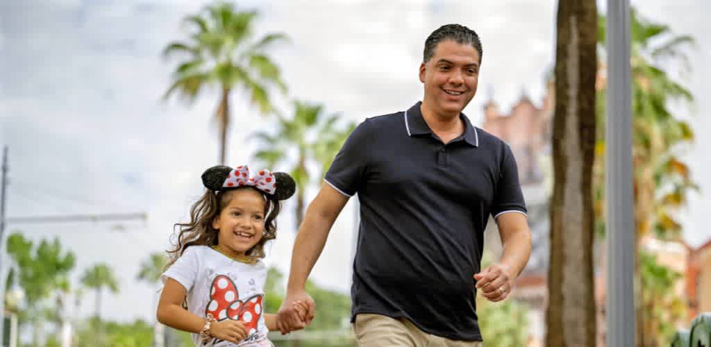 A man and a young girl are walking outdoors hand in hand, smiling, with palm trees and a sunny sky in the background.