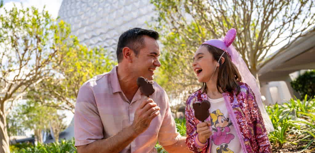 A smiling man and girl enjoy ice cream in a park, wearing fun costumes including a pink dress and bunny ears, surrounded by trees and greenery