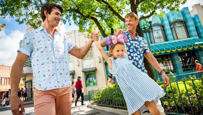 A joyful family strolls through Disney’s Hollywood Studios, with a young girl in Minnie Mouse ears playfully swinging between her parents amid the vibrant art deco architecture.